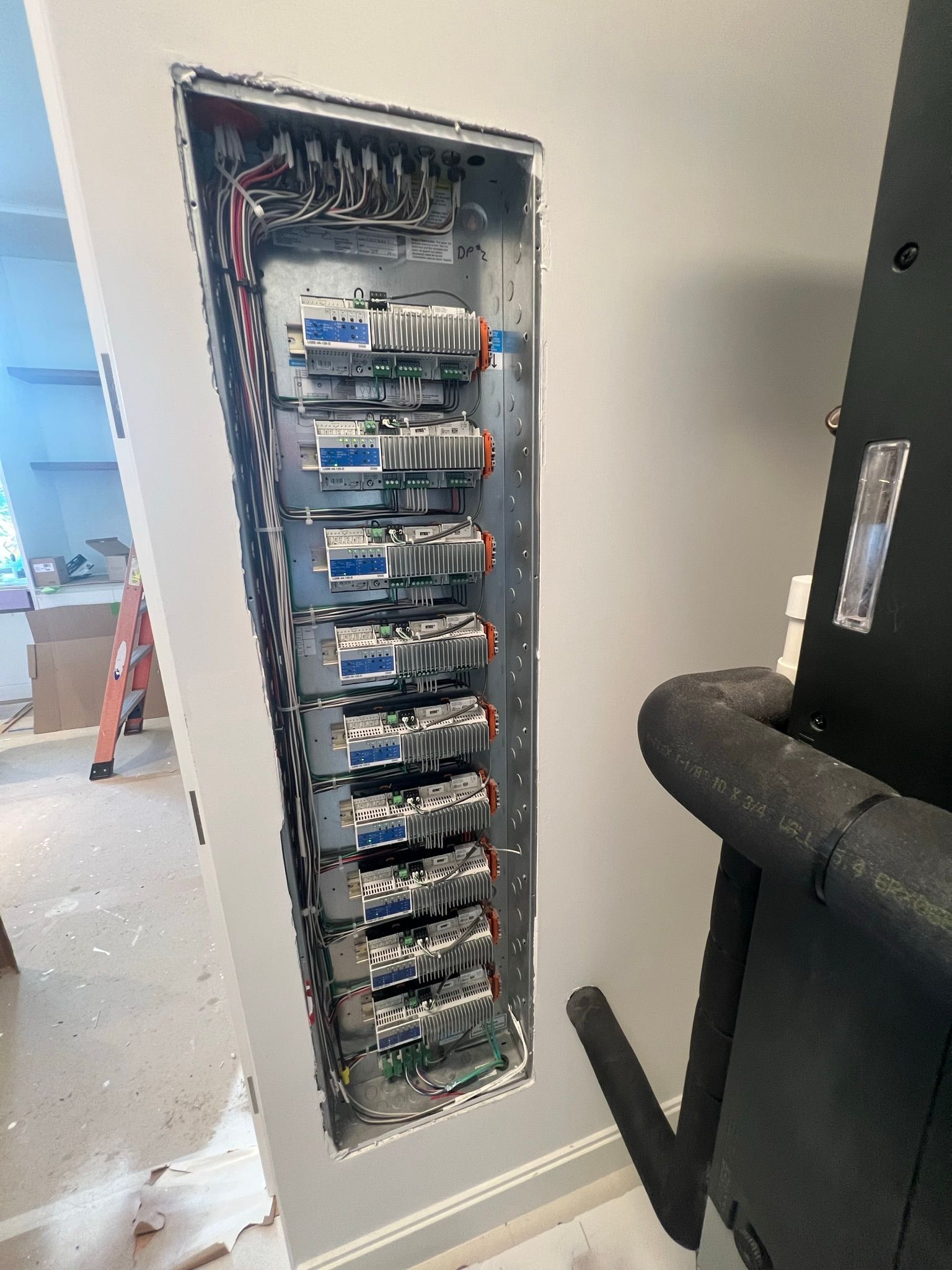 Open electrical panel recessed in a gray wall, wires and circuit breakers visible. Black pipe on right.