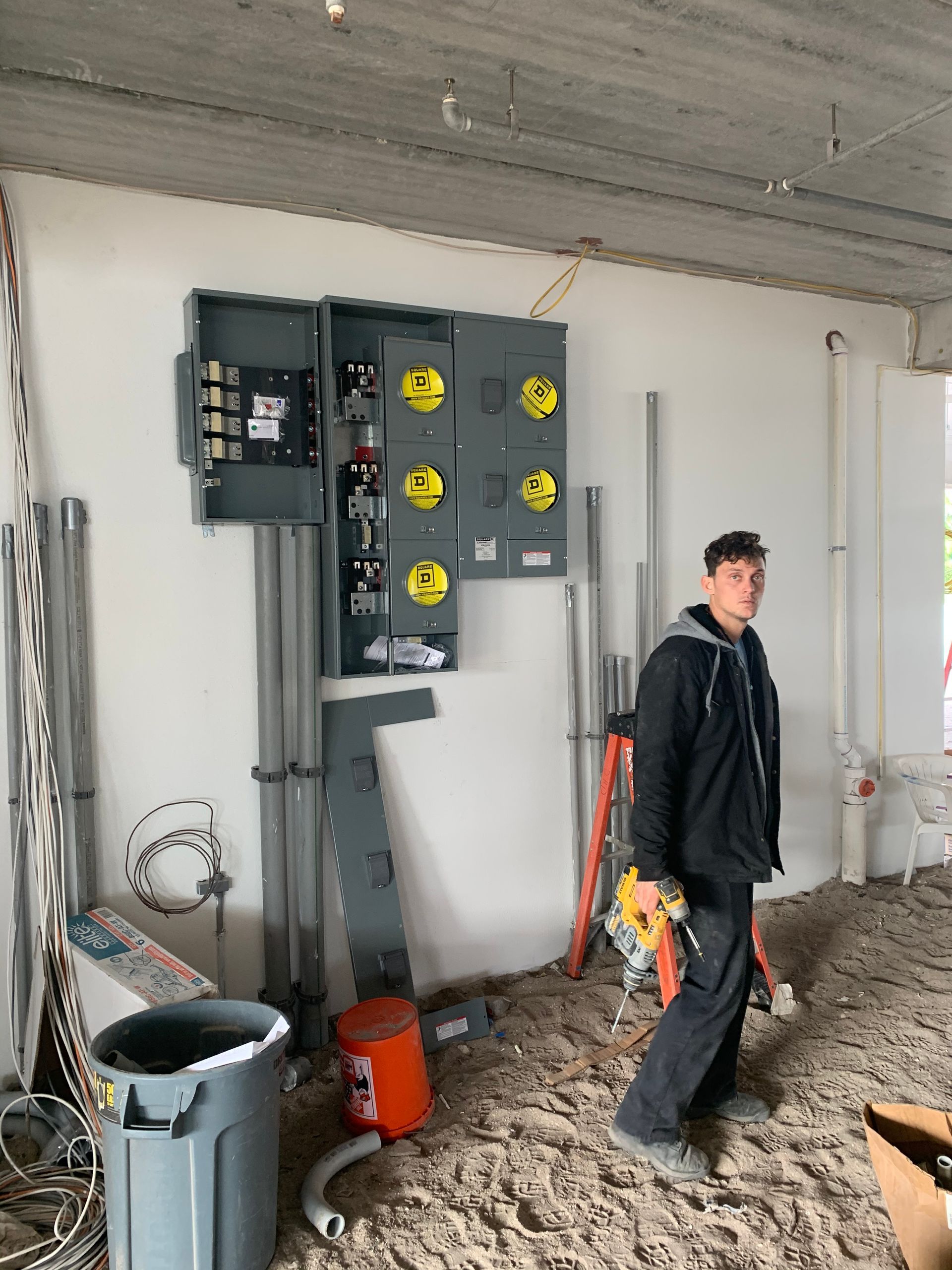 Electrician standing near electrical panel, construction site. Grey panel, conduit, and cables. Man in black jacket holding tools.