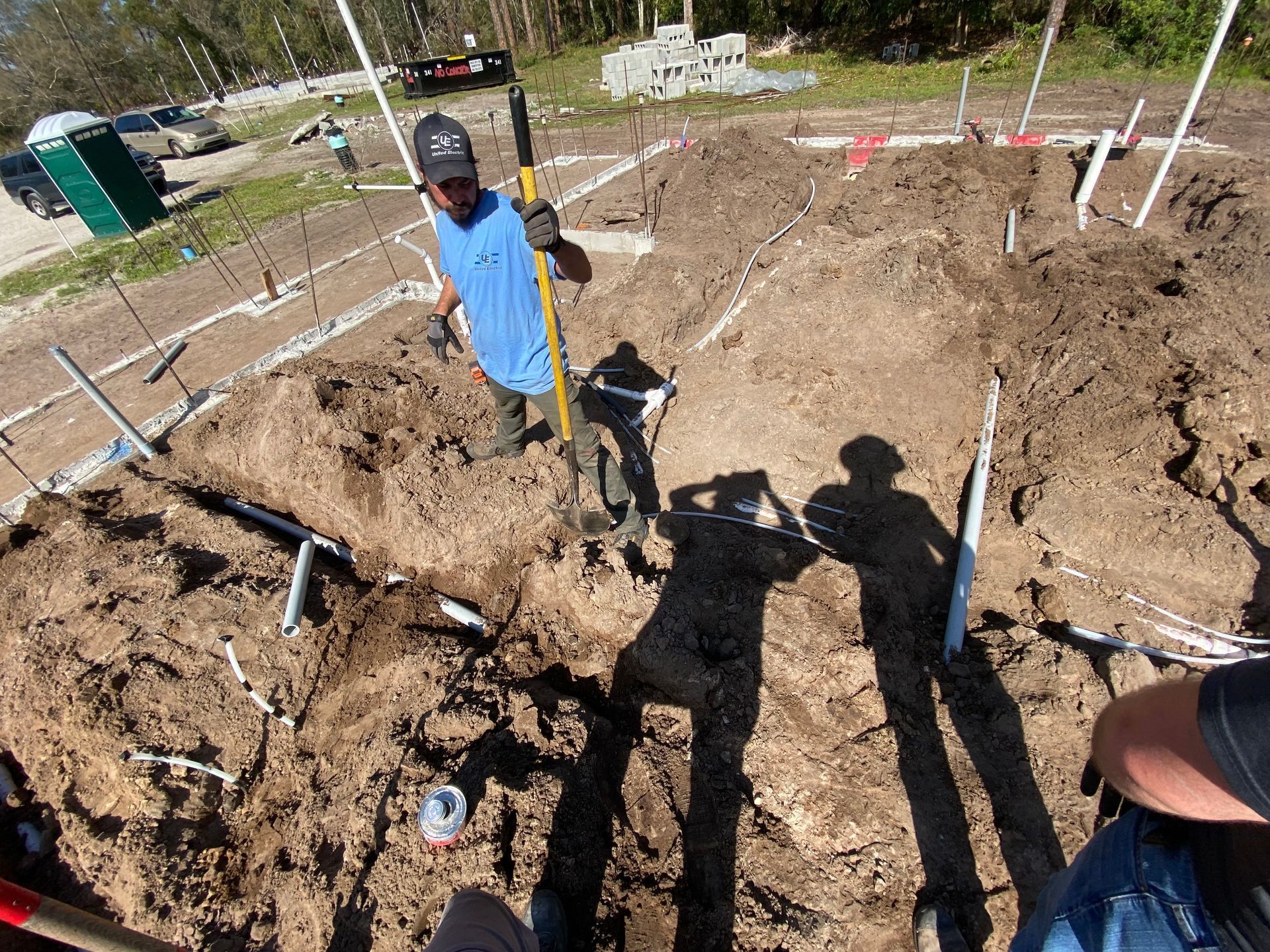 Man standing in dirt at a construction site, holding a tool. Plumbing and markers are visible.