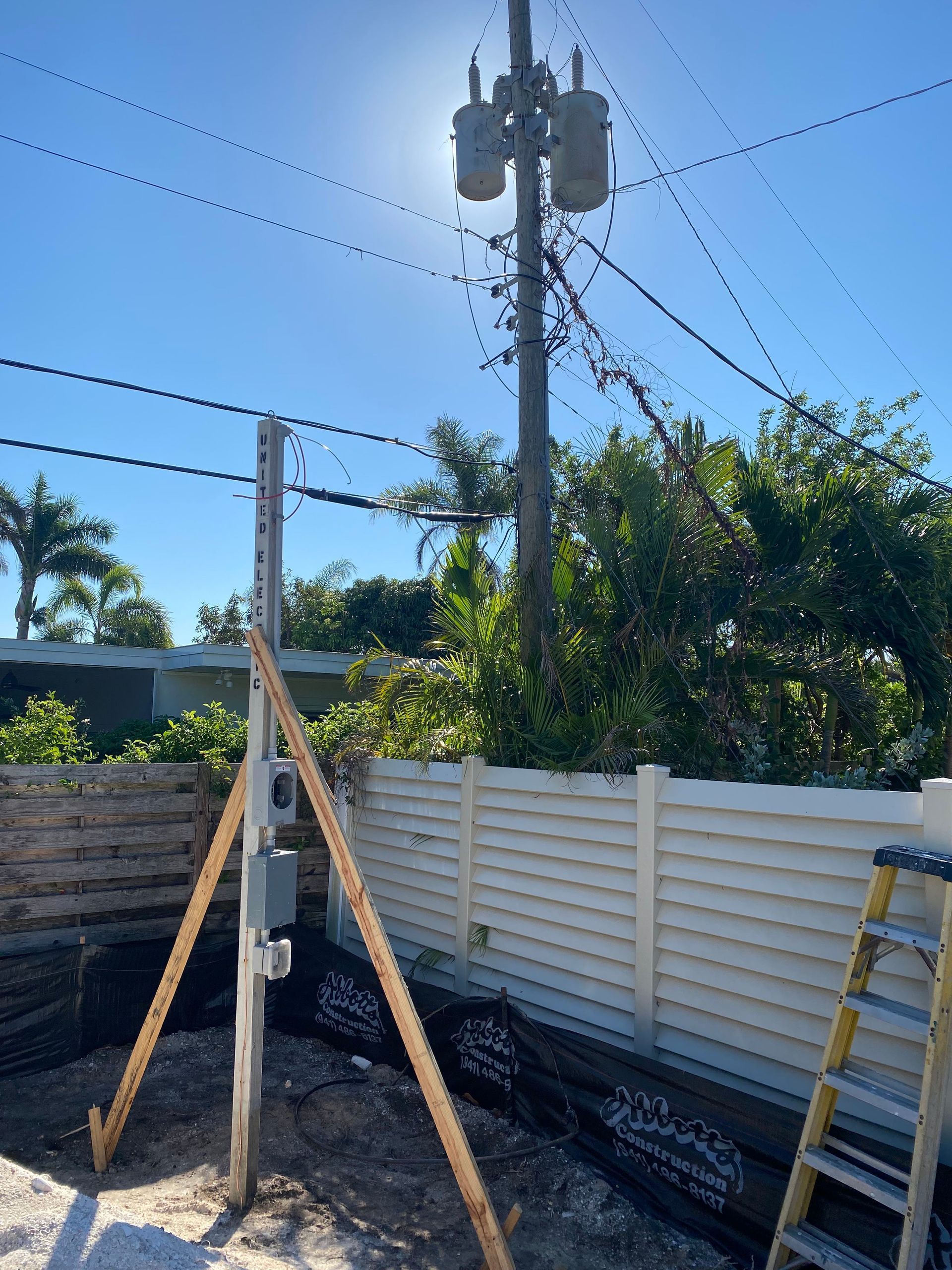 A utility pole with temporary supports next to a white fence under a sunny sky.