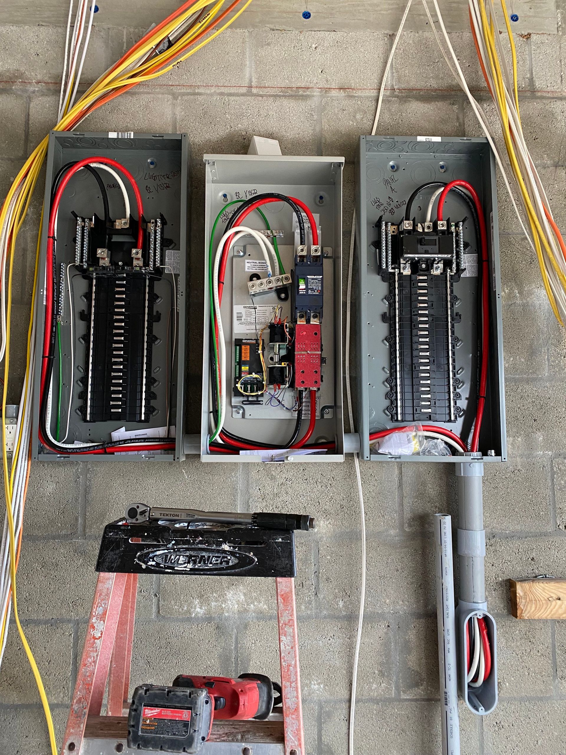 Three electrical panels on a wall with wiring, a red ladder, and tools.