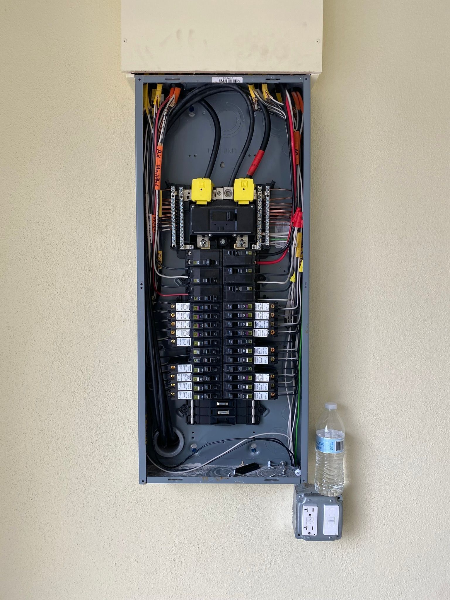 Open electrical panel on a wall with wires and breakers; water bottle and small gray box are next to it.