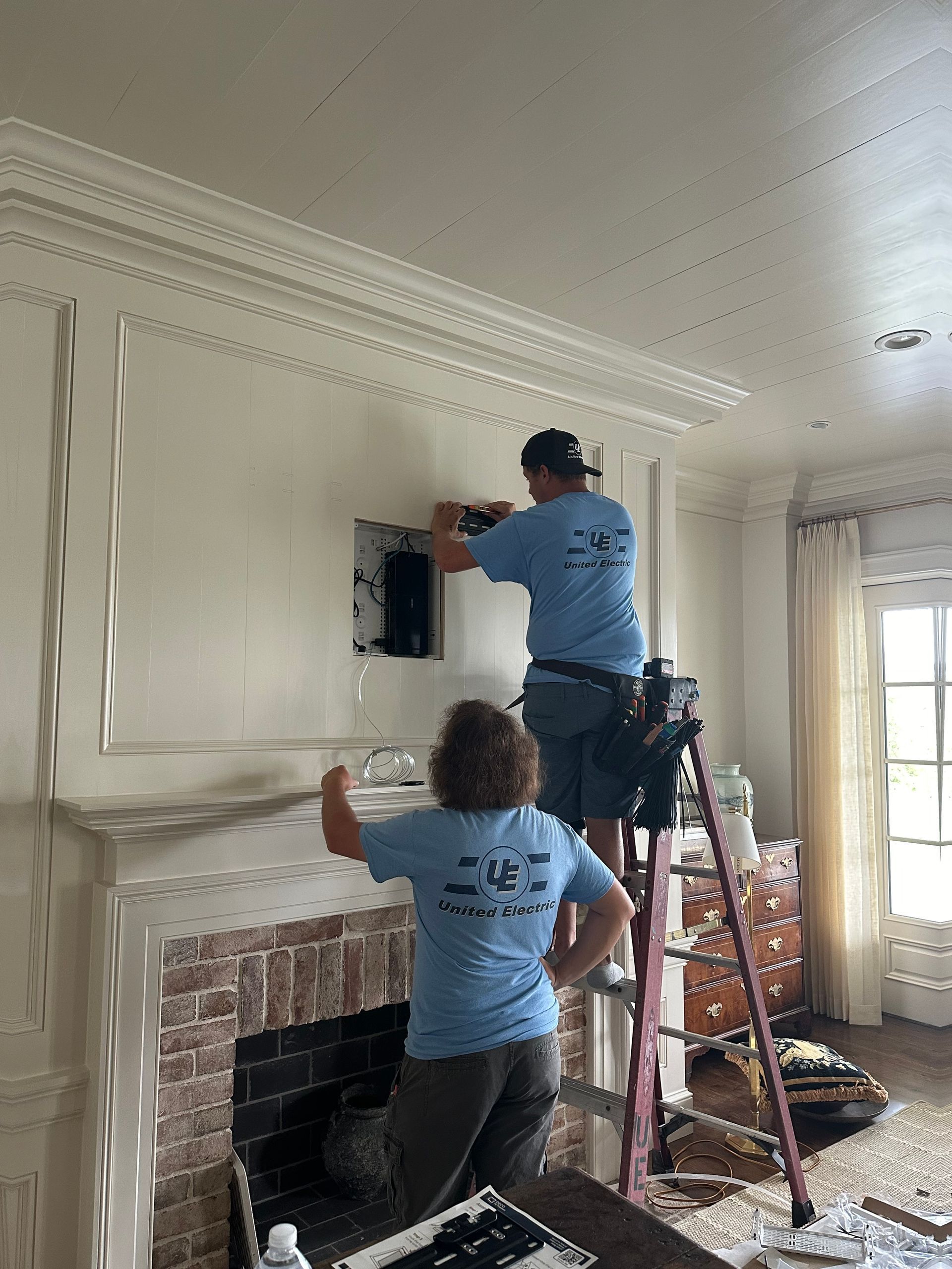 Two people installing electronics inside a wall above a brick fireplace, using a ladder.