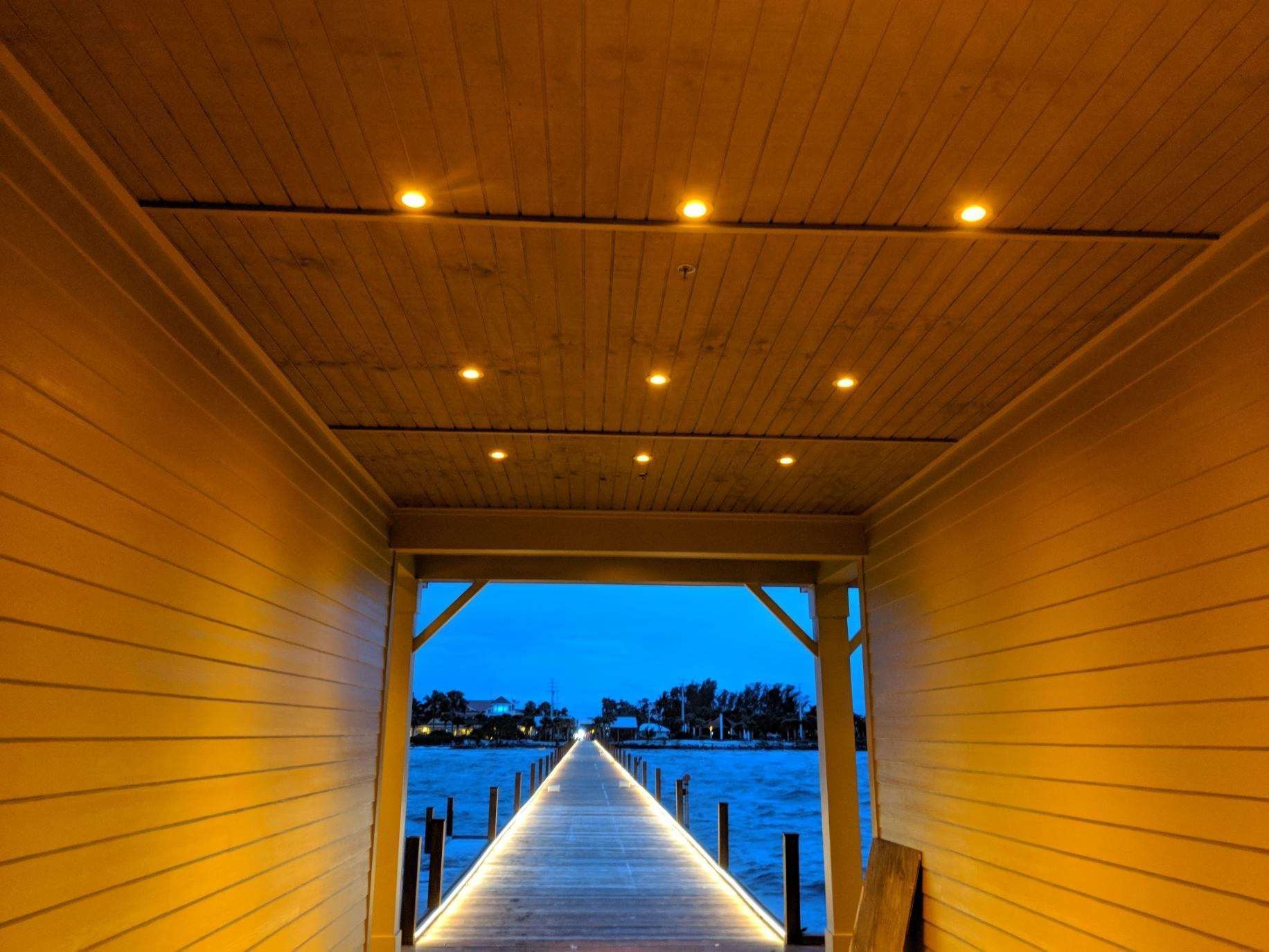 Wooden pier lit with lights, leading to a dark blue water with trees and sky at the end.