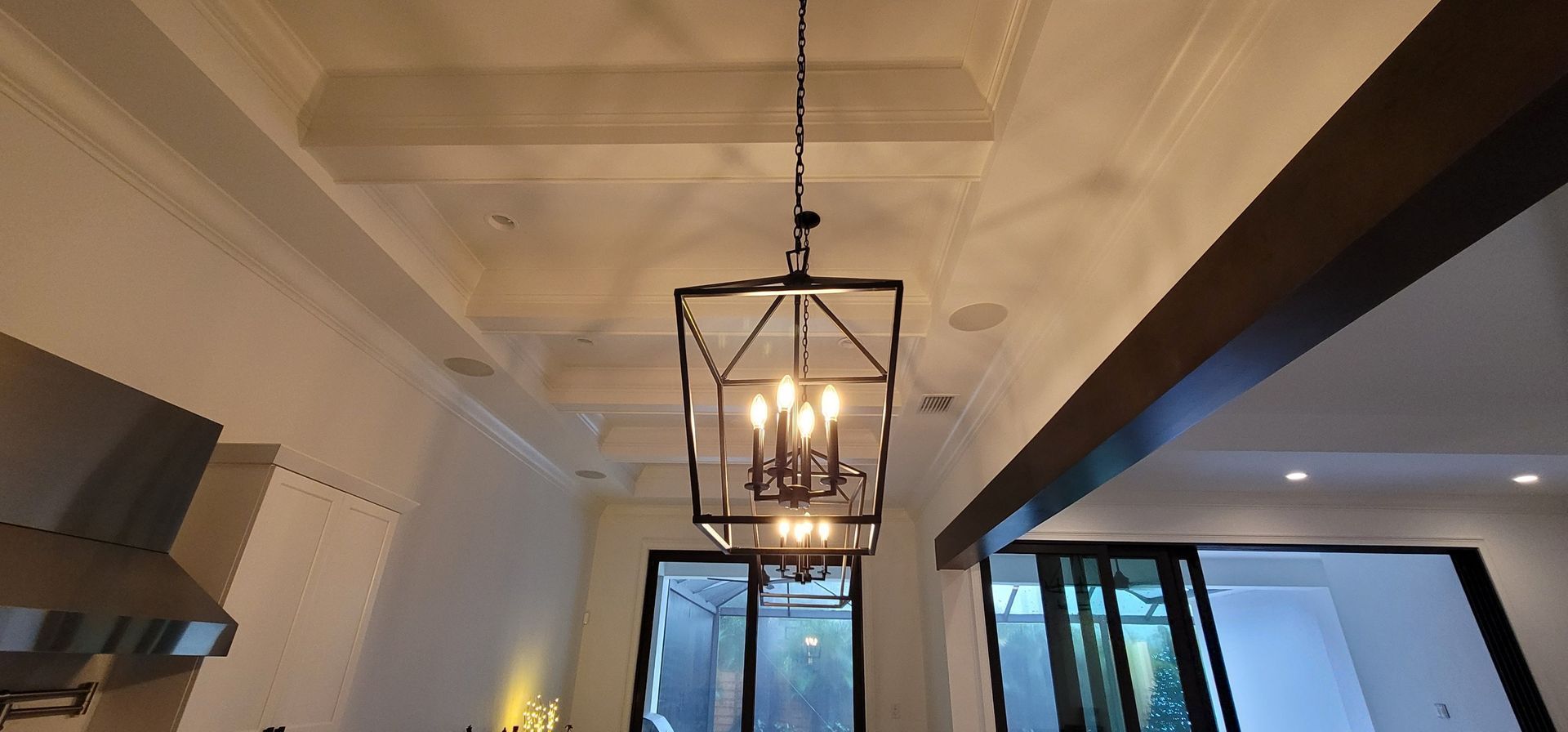 Ornate black chandelier hanging from a white coffered ceiling. Brown beams and glass door visible.