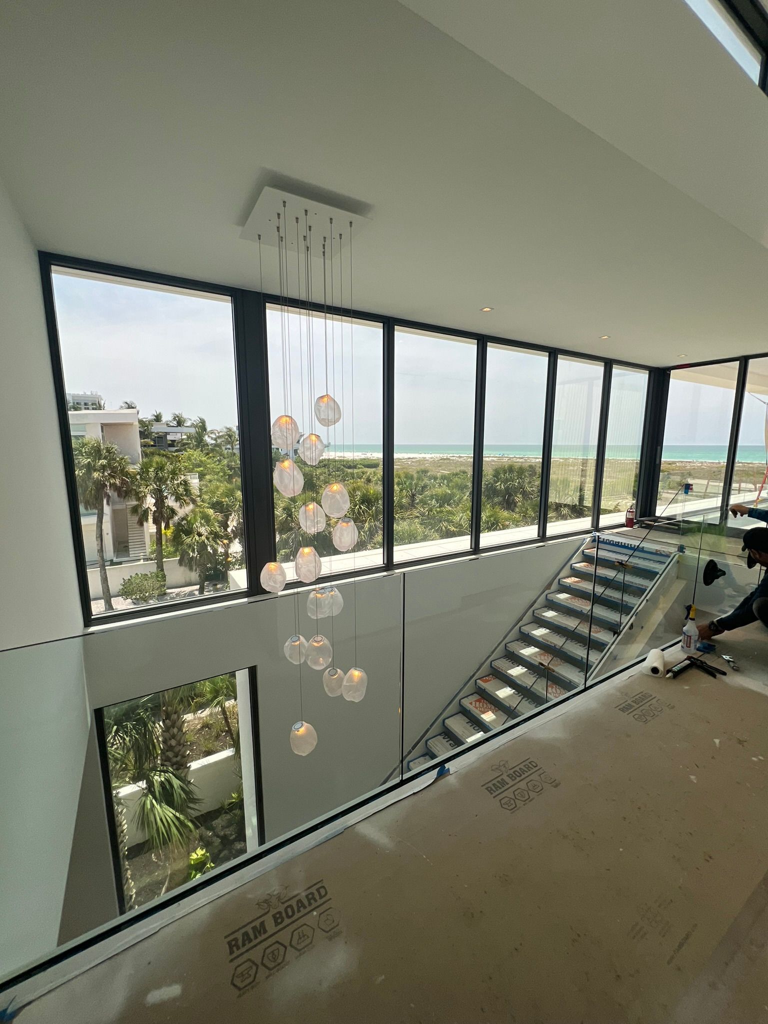 Modern house interior with staircase, large windows overlooking beach and ocean; chandelier hangs.