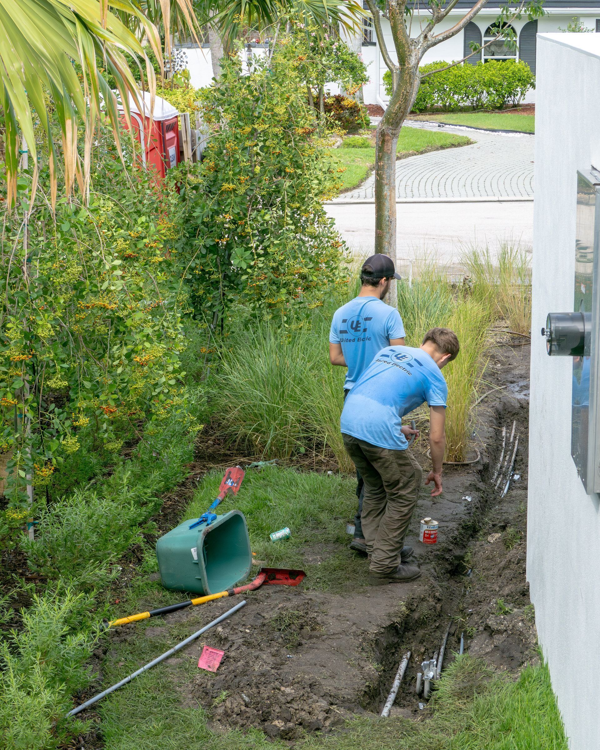 Two workers in blue shirts are digging in a yard near a house. Green plants surround the work area.