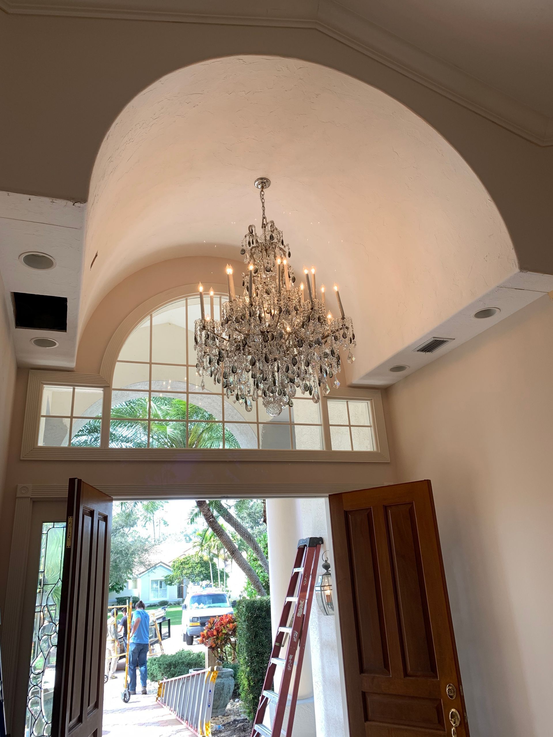 Entryway with large chandelier under arched ceiling, above a window with greenery visible through it.