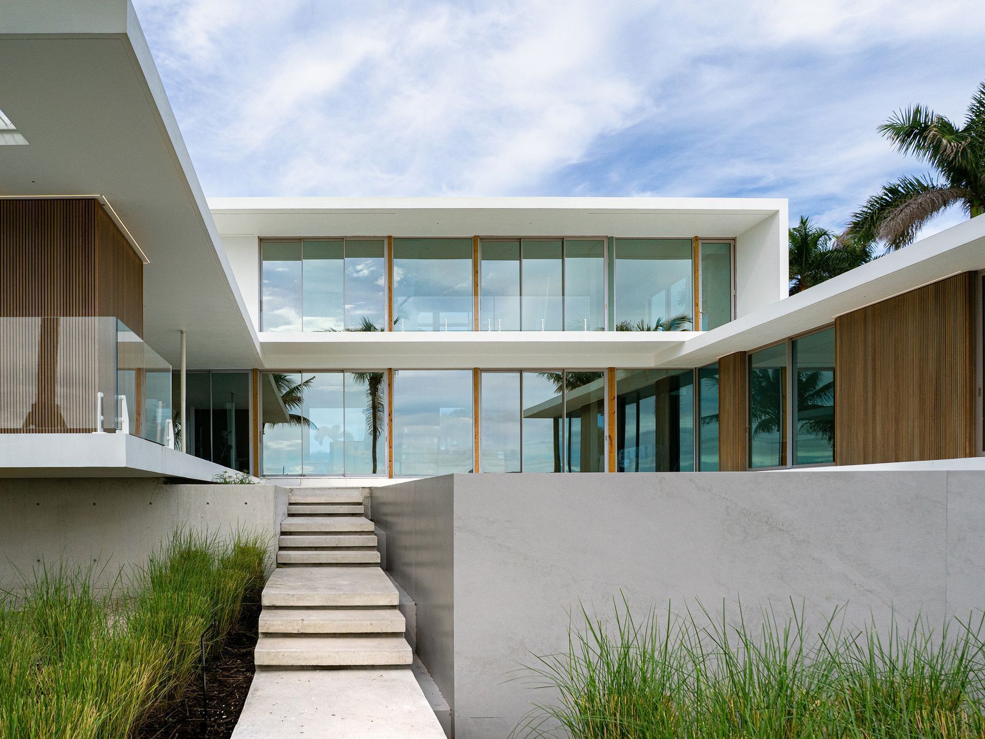 Modern white house with glass walls and a staircase leading up. Palm trees and cloudy sky in background.
