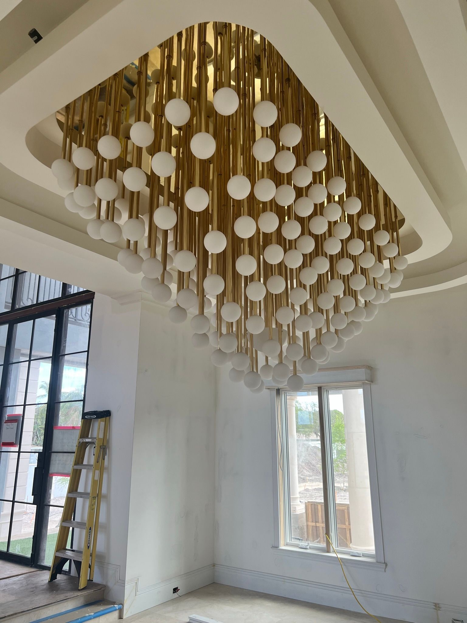 Gold and white chandelier hanging in a room with a window, ladder, and white walls.
