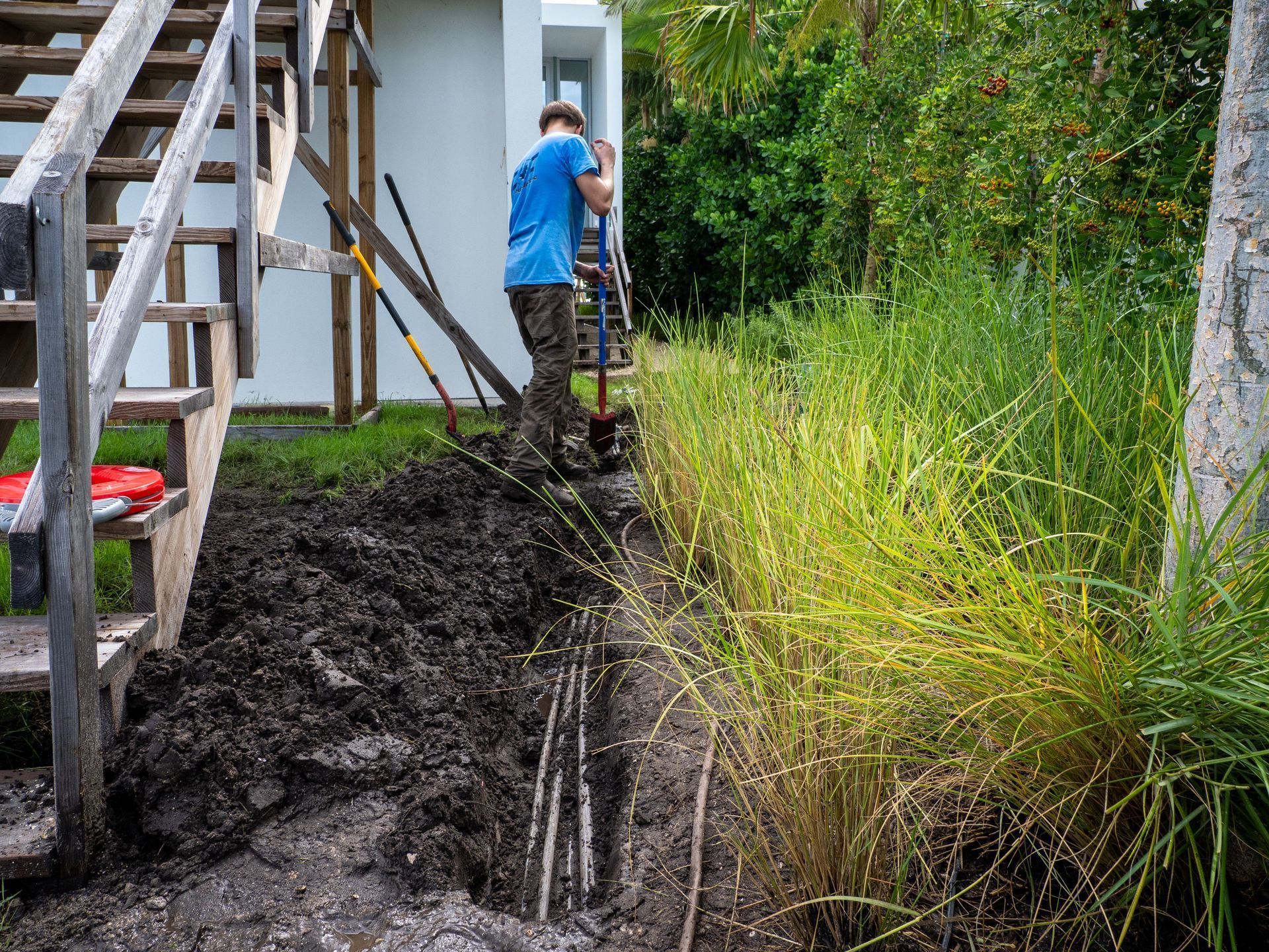 Man digging trench next to tall grasses near a house with wooden stairs.