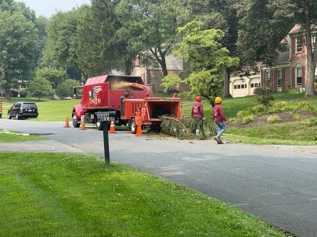 Red utility truck and workers repairing a suburban road beside a grassy lawn and trees.