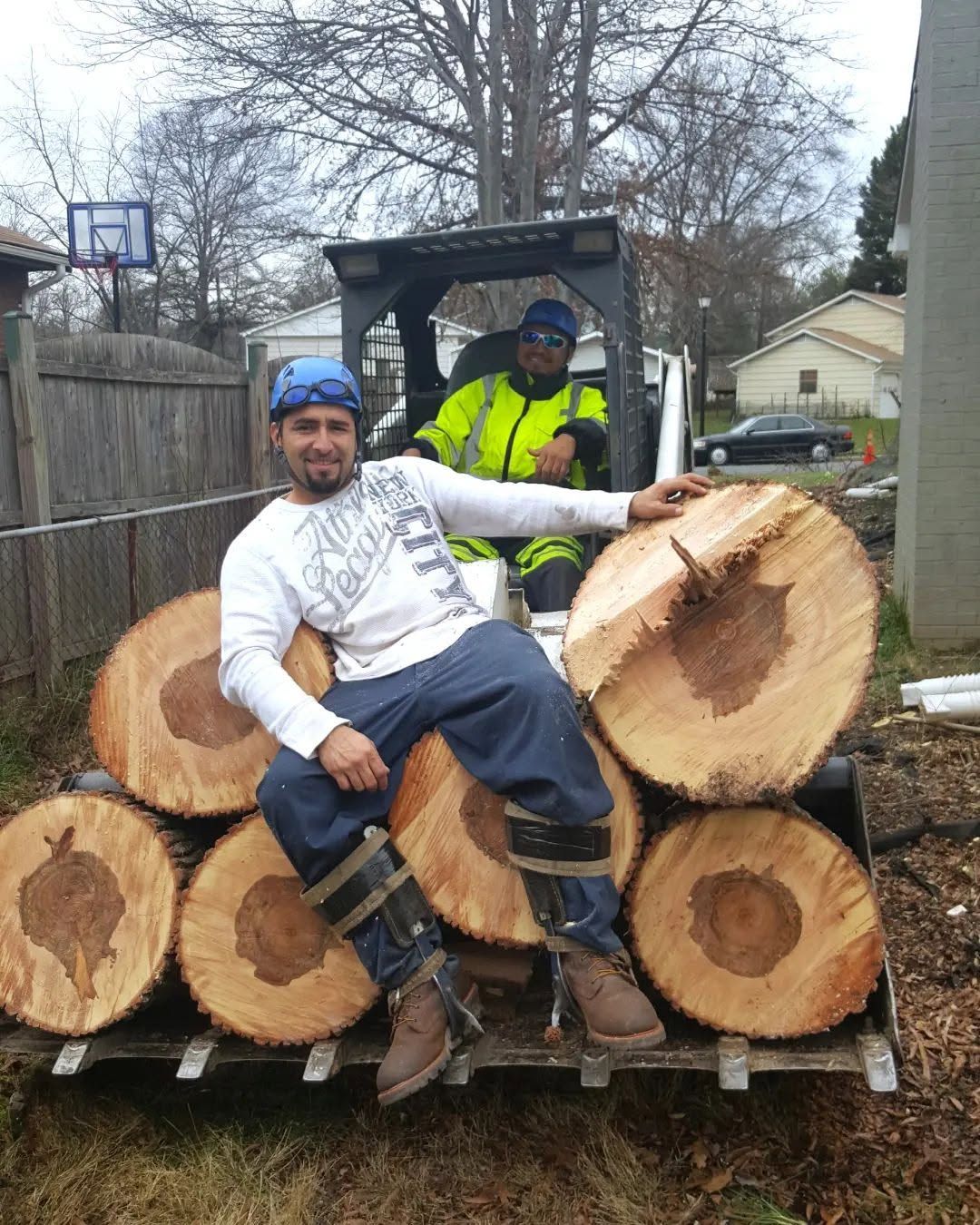 Two men on a trailer piled with cut logs, one sitting in front of a loader in a backyard.