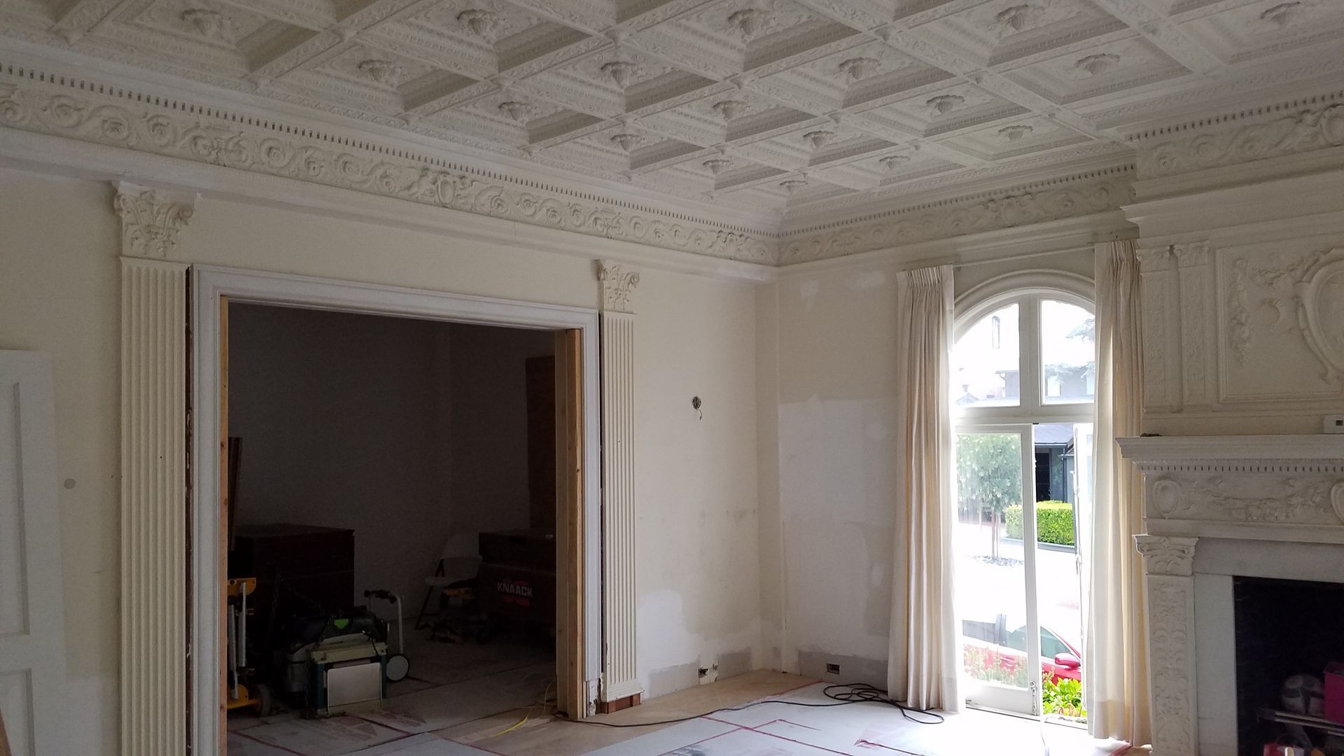 Ornate ceiling and fireplace in a room with two large windows and beige curtains; construction in progress.