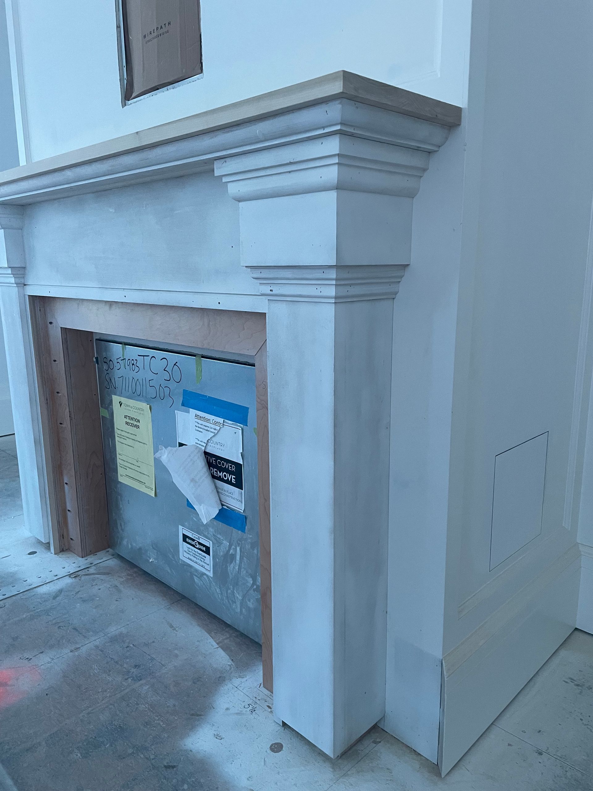 Gray painted fireplace with a light-colored wood mantel against a white wall; unfinished construction.
