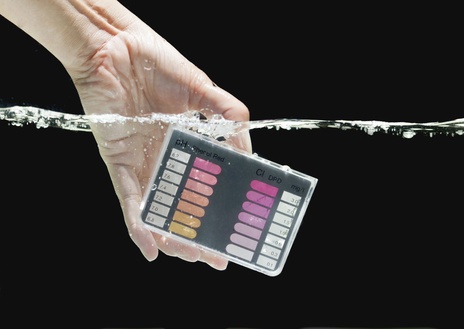 A hand holds a pH test strip chart partially submerged in water against a black background.