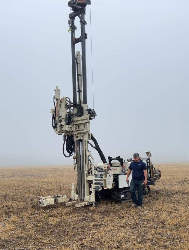 Worker beside a large drilling rig in a barren field under an overcast sky