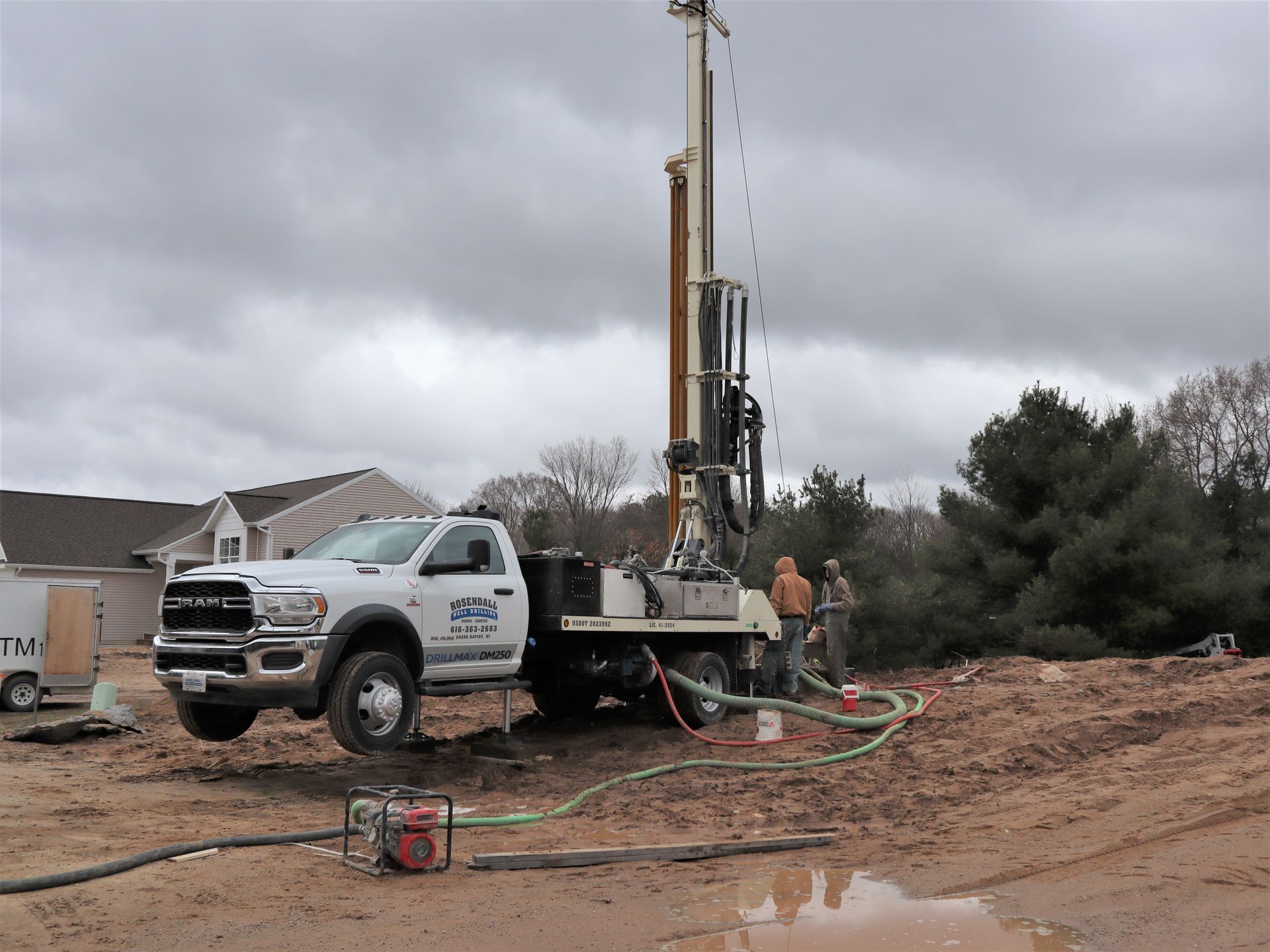 A white truck-mounted drill rig on a muddy construction site with two workers nearby under a cloudy sky.
