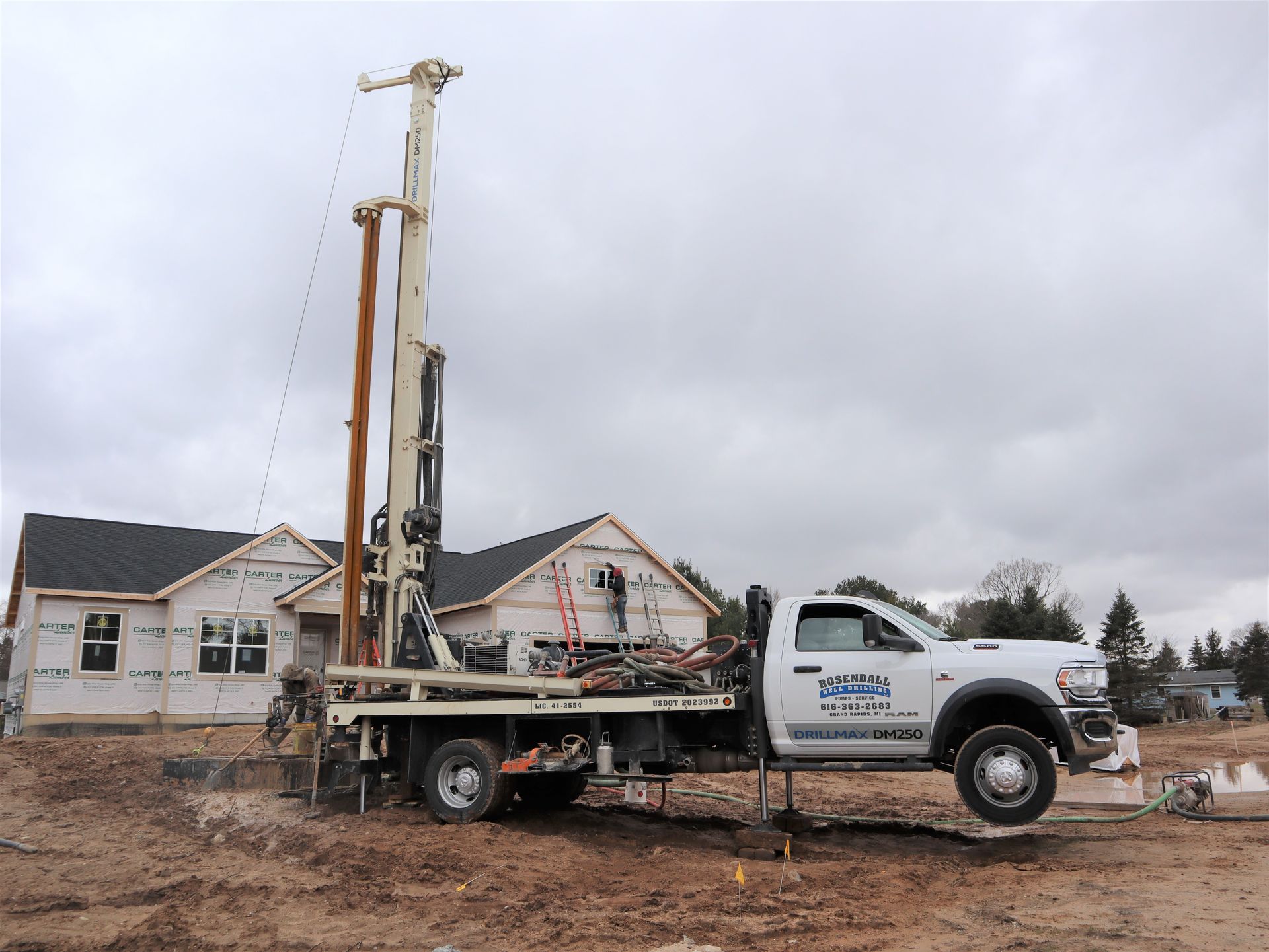A white drilling truck parked on a muddy construction site next to a partially built residential house.