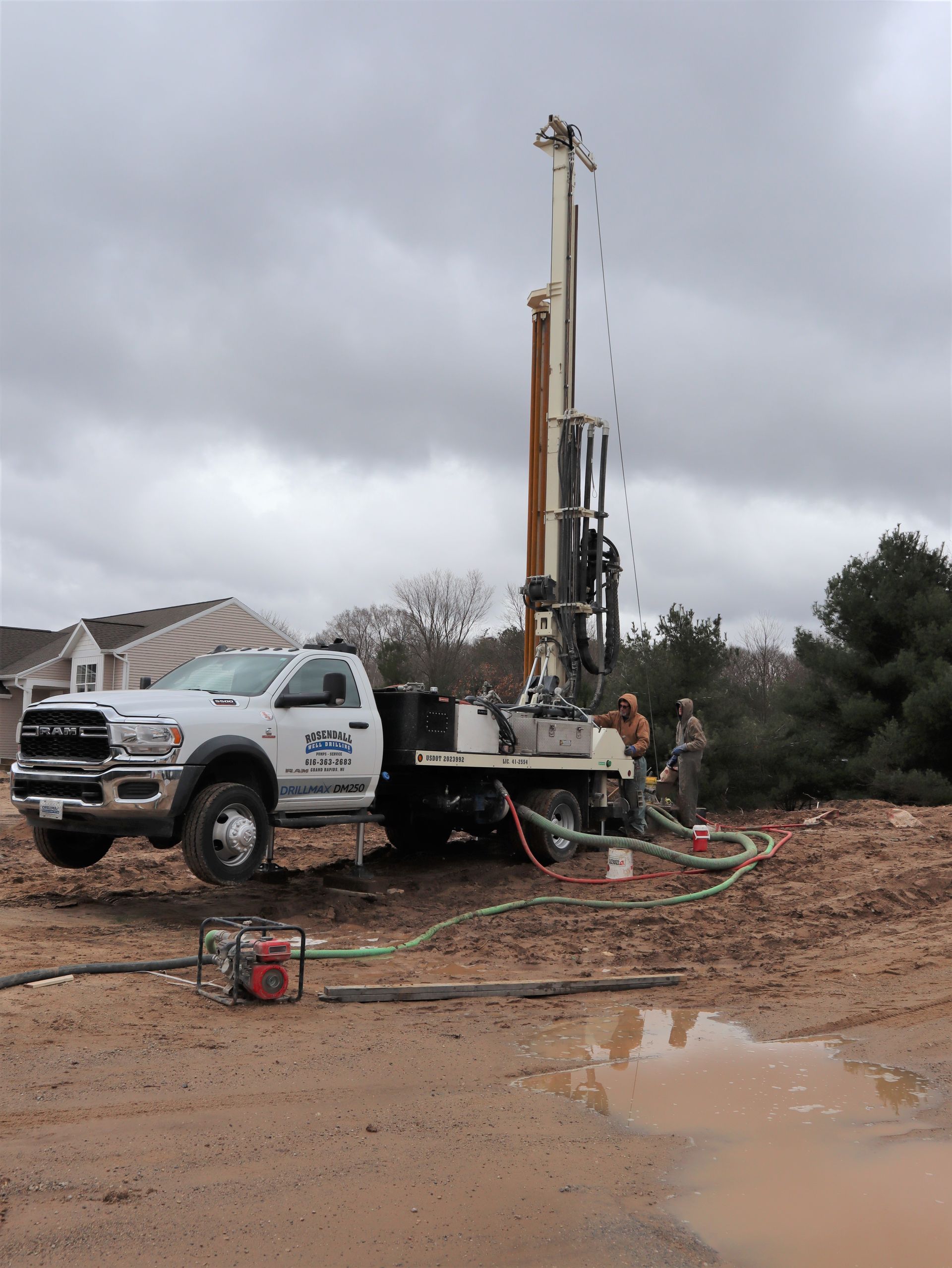 A white drilling rig truck operates in a muddy field near a house under a cloudy sky.
