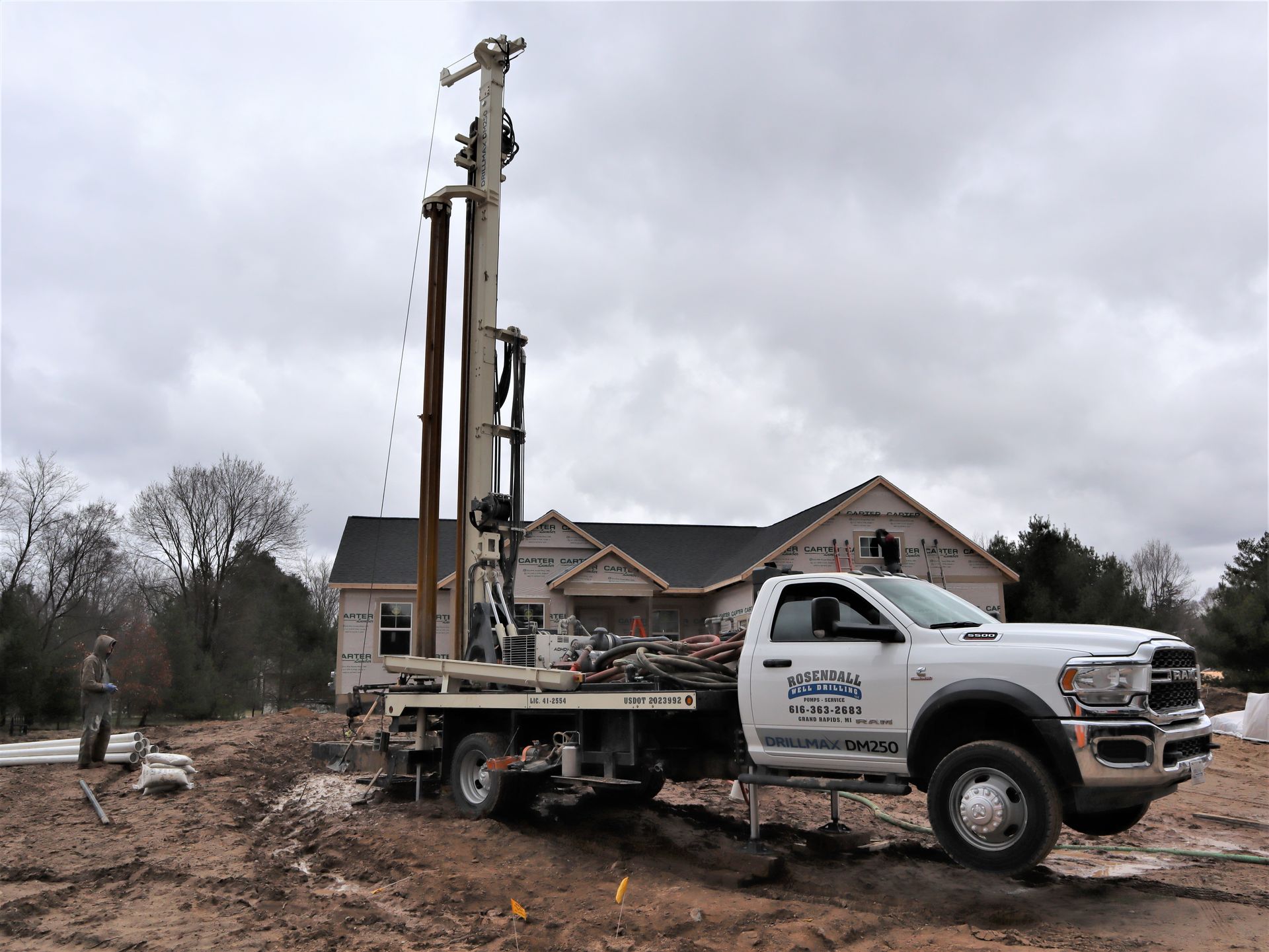 A white truck with a mounted drilling rig parked on a muddy construction site in front of an unfinished house.