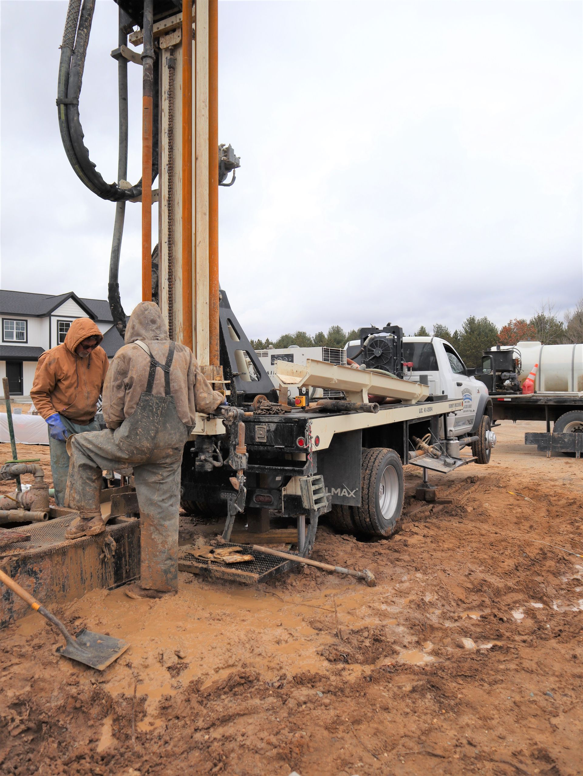 Two workers in work gear operate a truck-mounted drilling rig at a muddy, outdoor construction site.