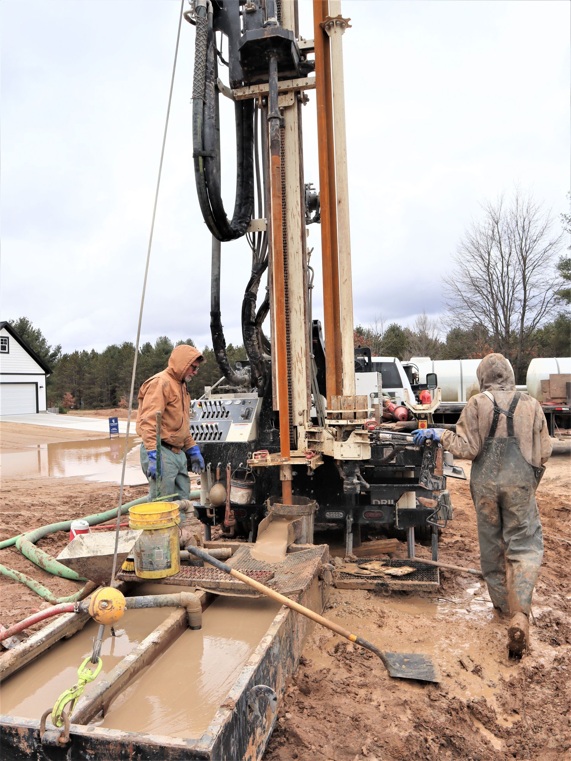 Two workers operate a tall drill rig on a muddy construction site under an overcast sky.