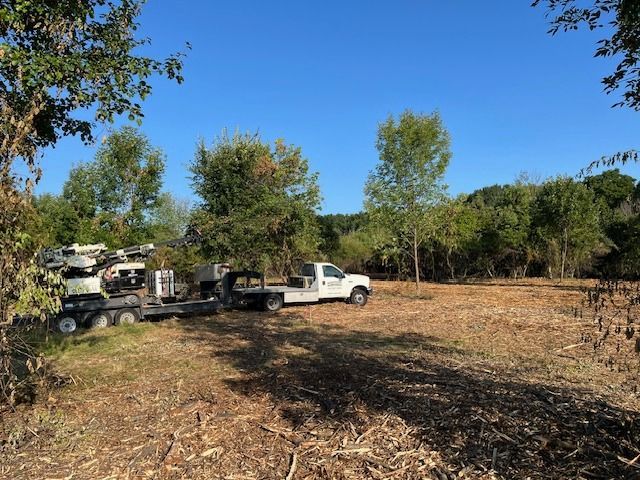 A white work truck with a trailer and heavy equipment parked in a partially cleared, wooded lot on a sunny day.