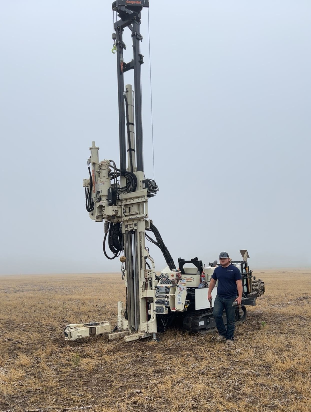 A person stands next to a large, white industrial drilling rig positioned in a vast, flat field under a foggy sky.