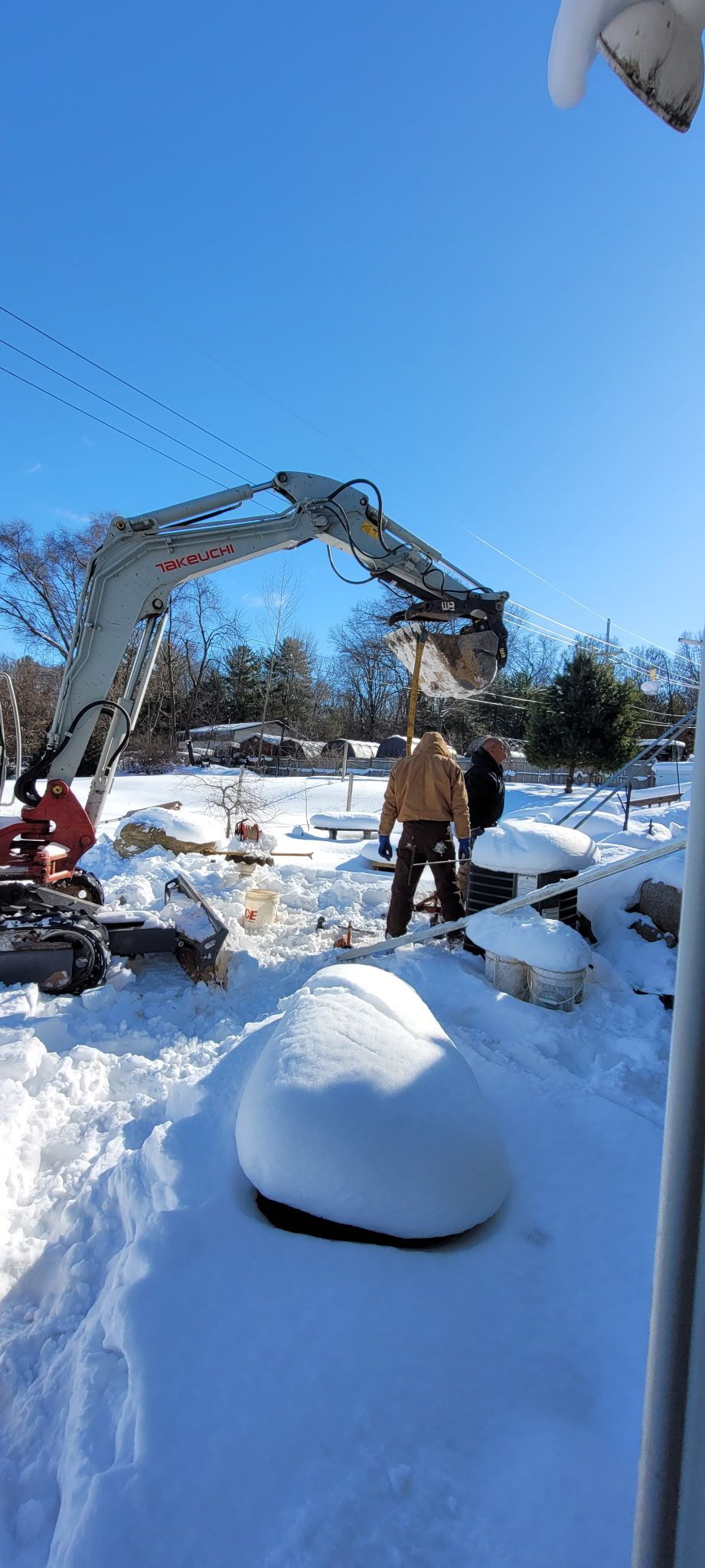 An excavator moves snow in a bright, sunny yard blanketed in deep snow, with two people working near the machine.