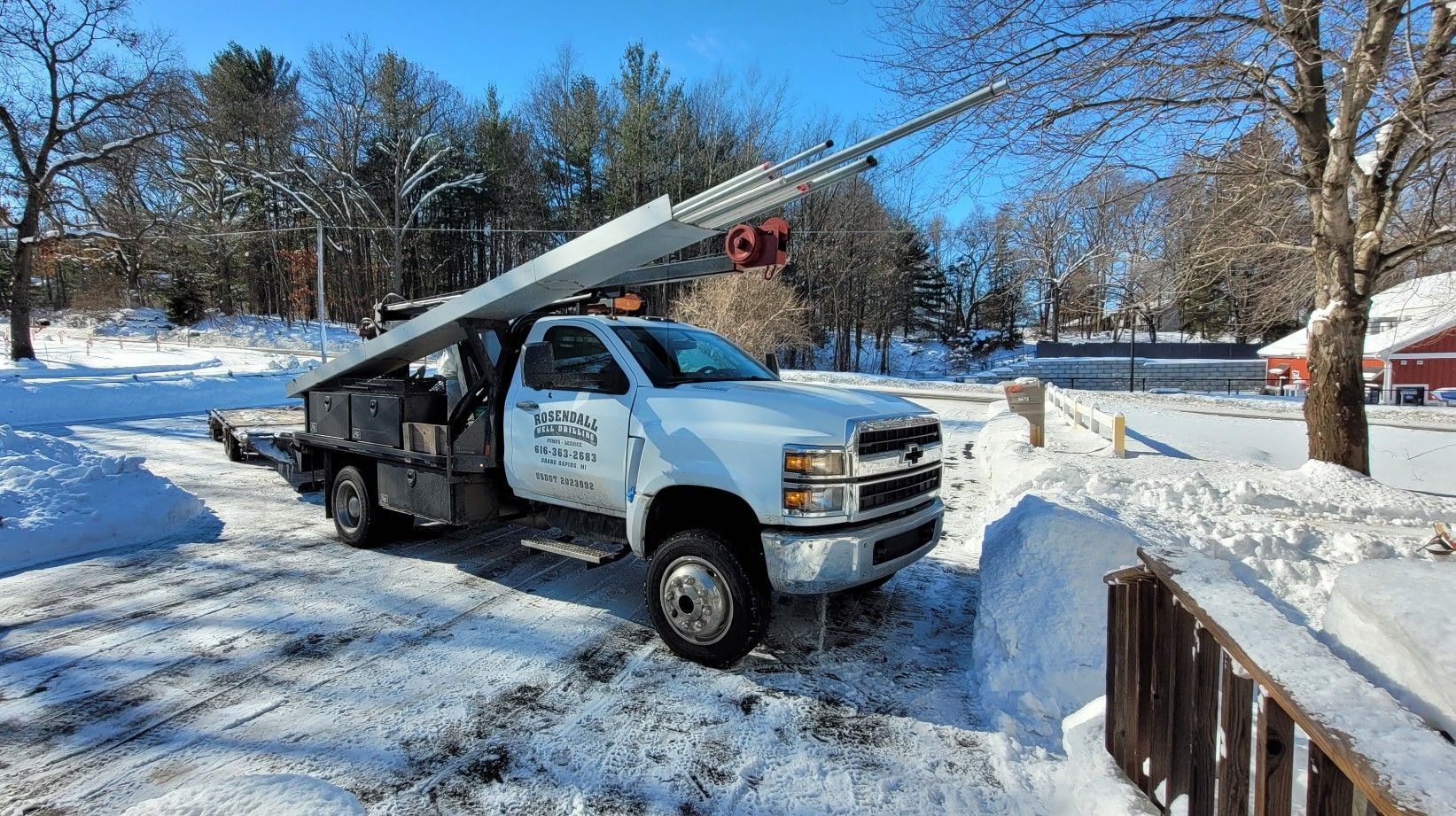 A white utility truck with a long ladder attached to its bed parked on a snowy, sunny landscape.
