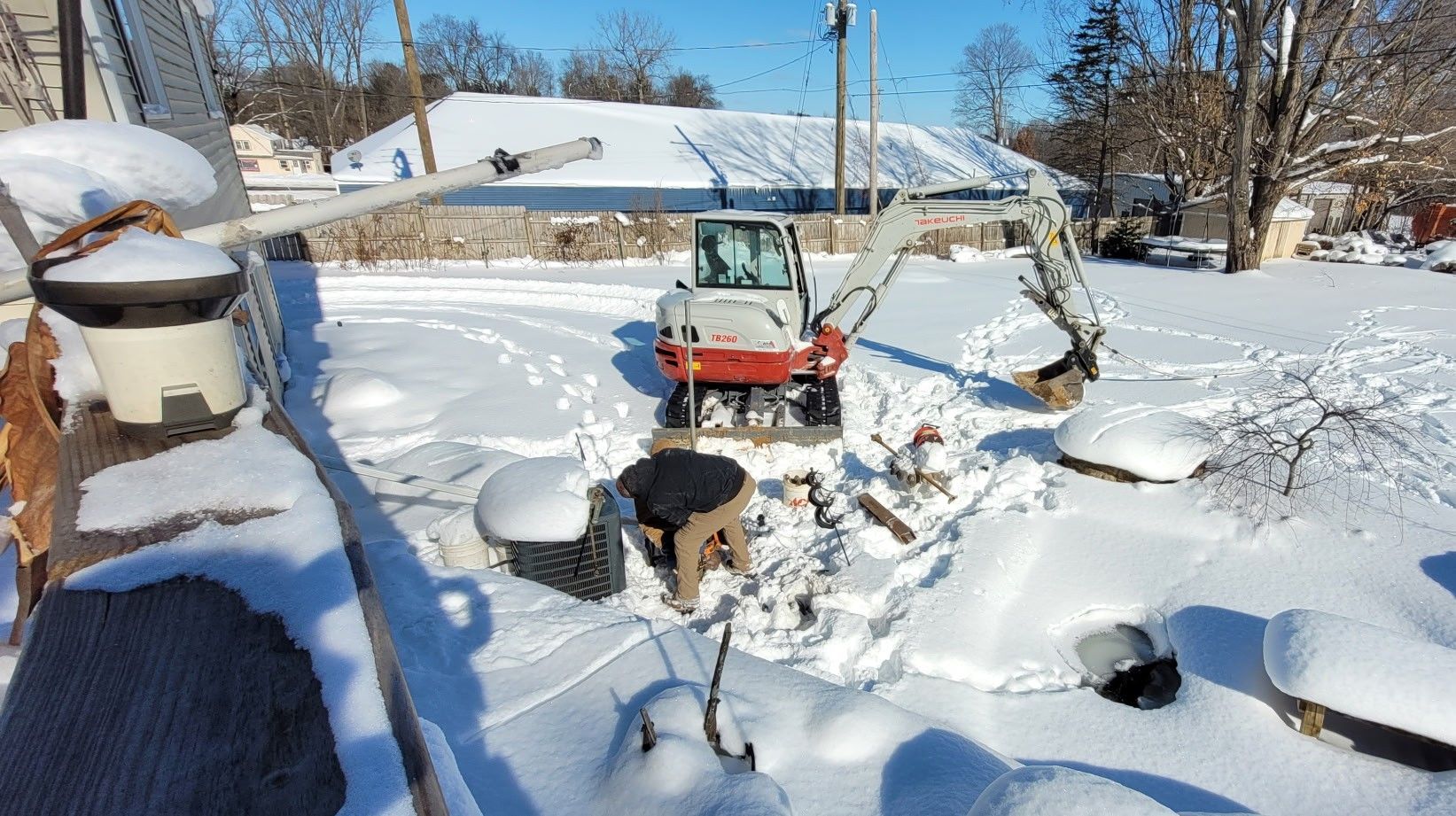A person operates a small construction excavator in a snowy yard while clearing ground for a project.