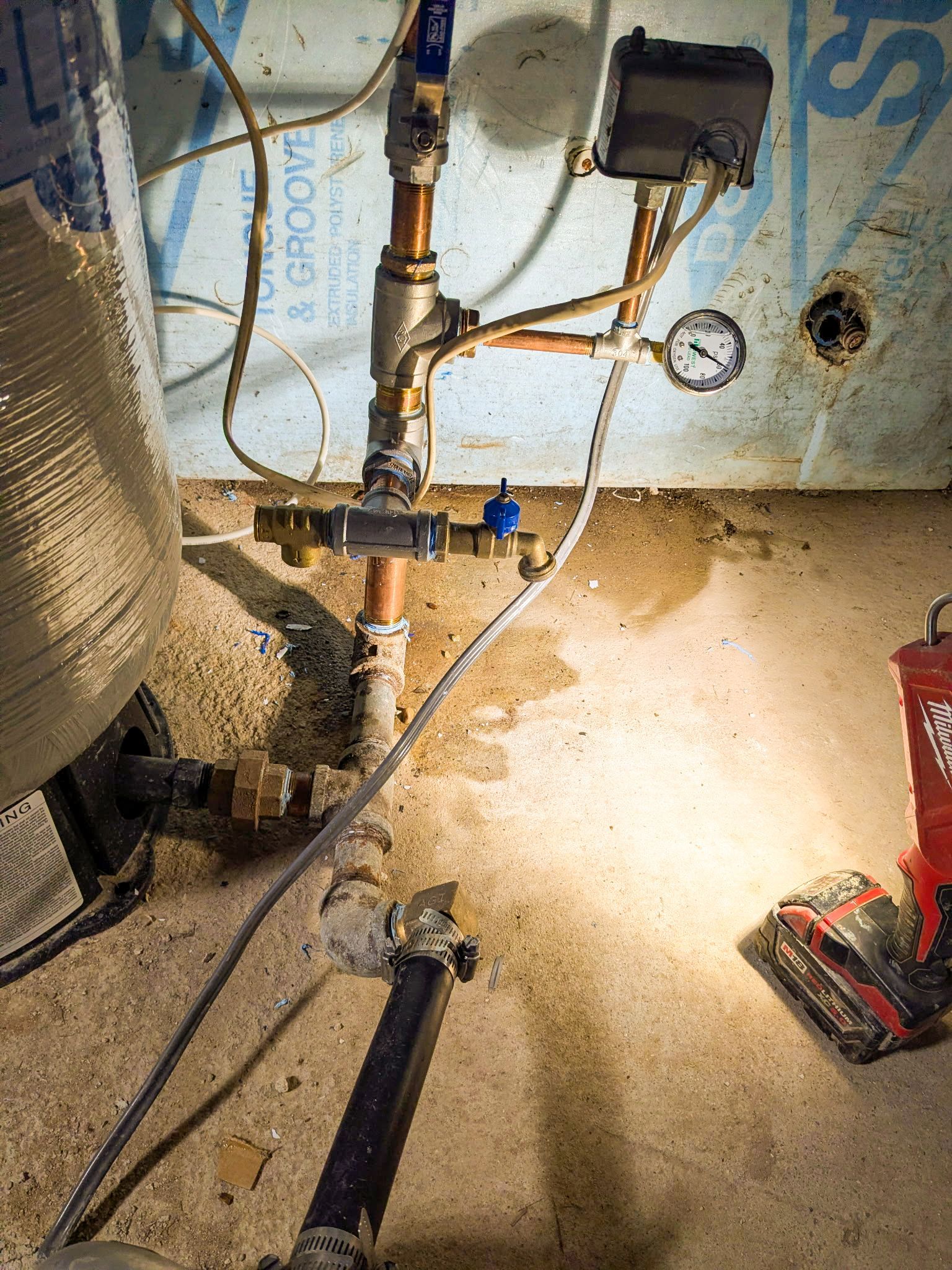 A water well pressure tank setup with copper pipes, a pressure gauge, and a black electrical control switch on a wall.
