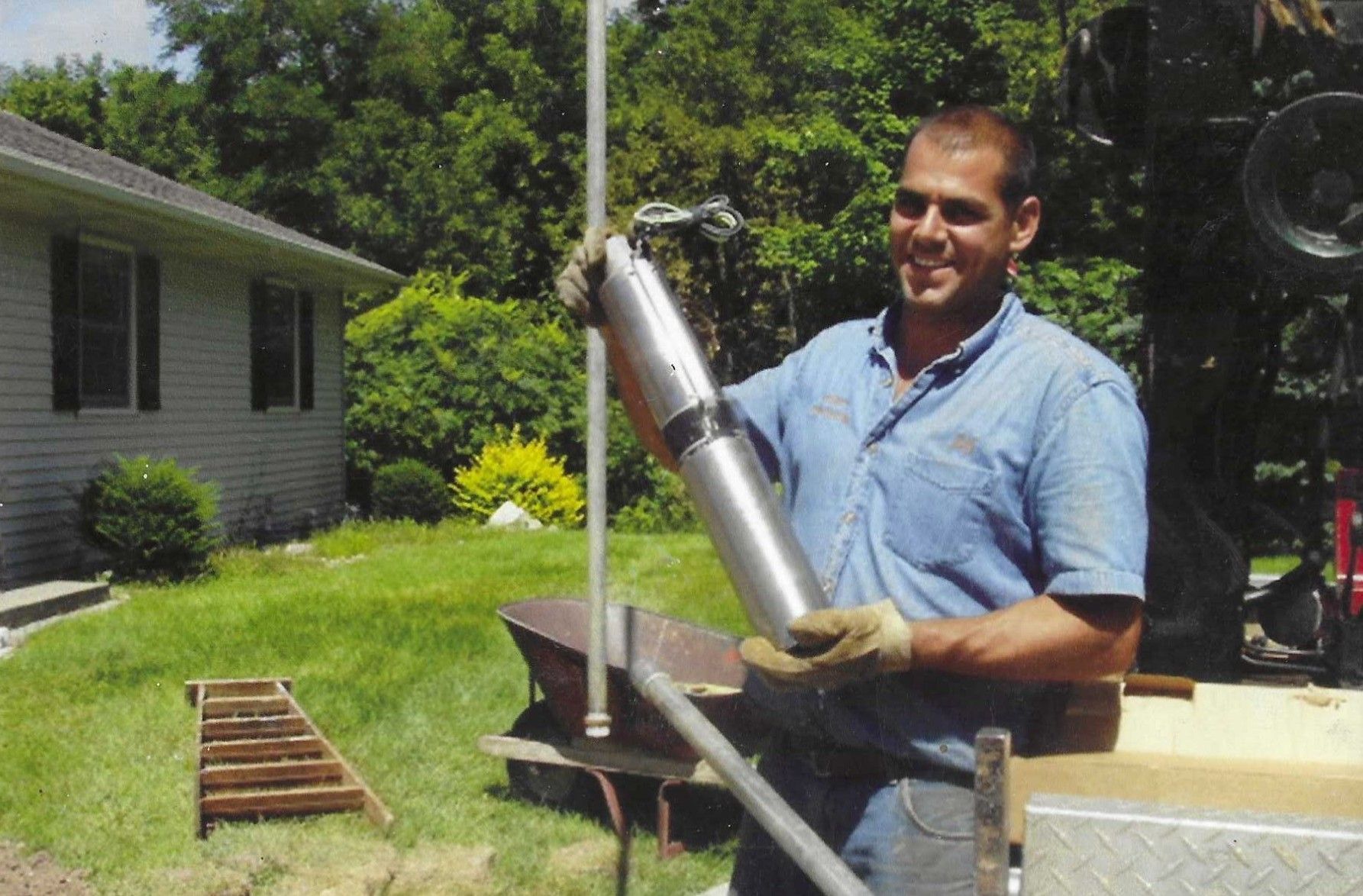 A smiling technician holds a stainless-steel submersible water pump outside a residential home.
