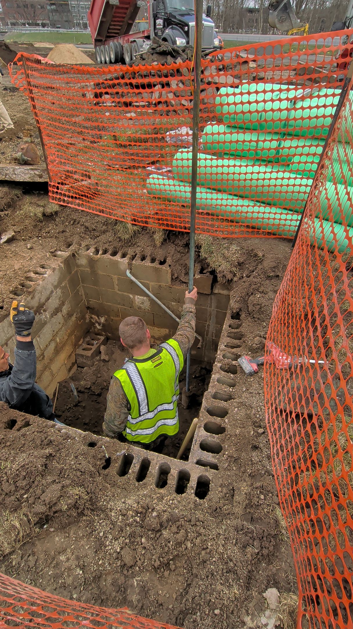 A construction worker in a high-visibility vest stands in a square, excavated trench near stacked green drainage pipes.
