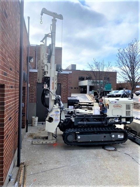 Tracked drilling rig beside a brick building on a concrete lot under a cloudy sky