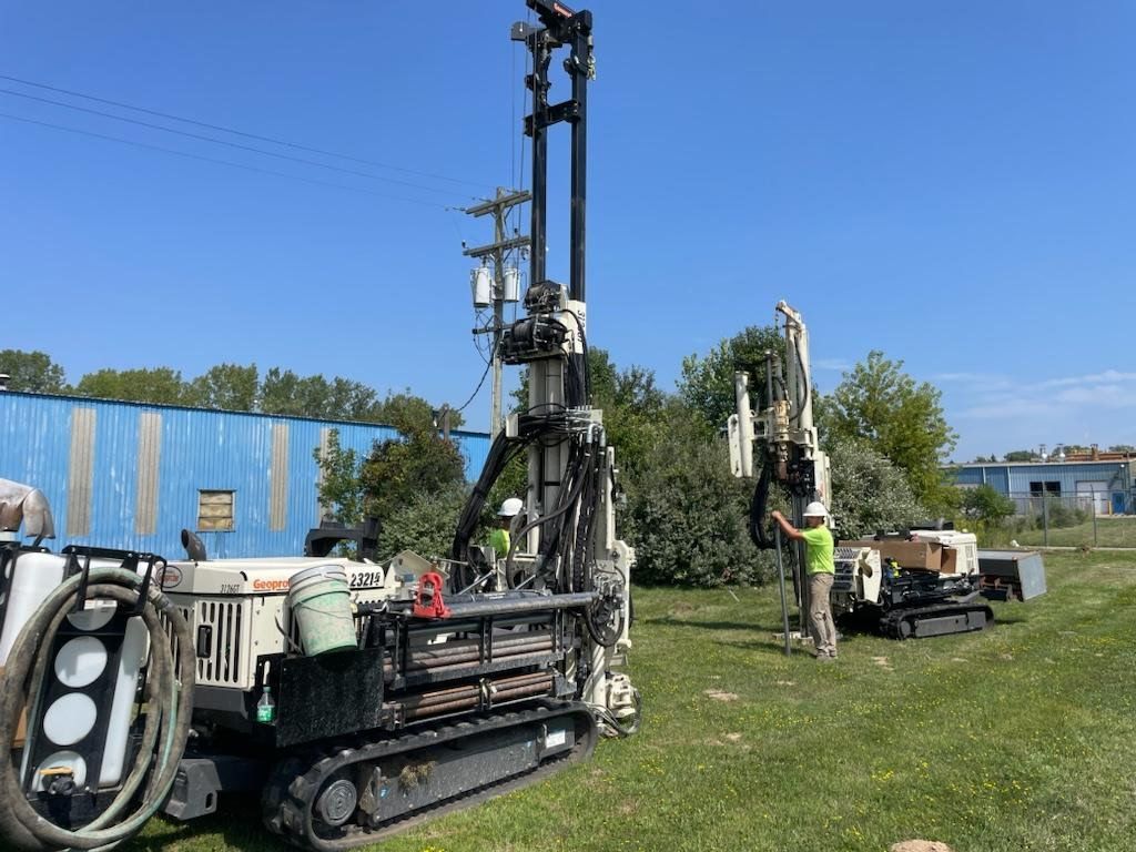 Workers beside a large drilling rig on a grassy site under a blue sky
