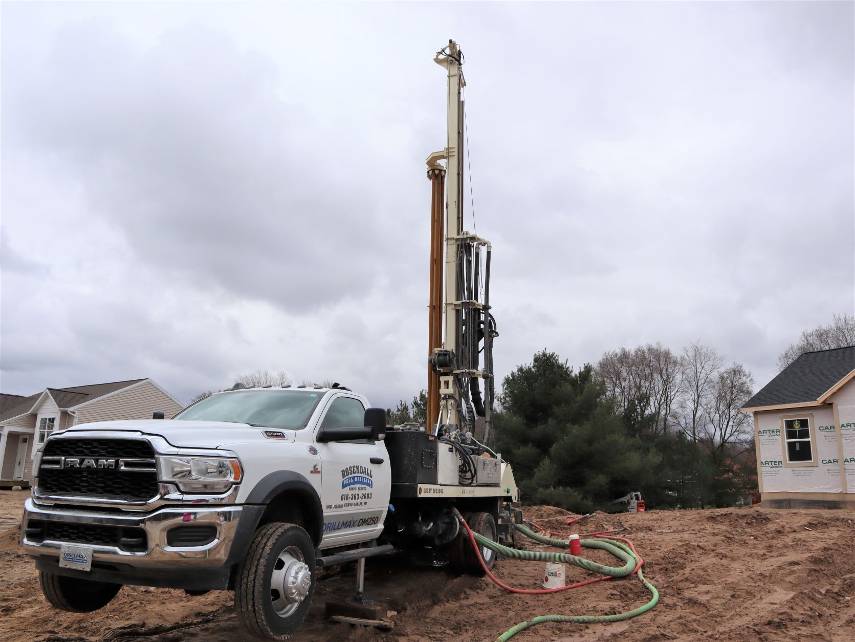 White drilling truck with tall rig on a muddy construction site under cloudy skies