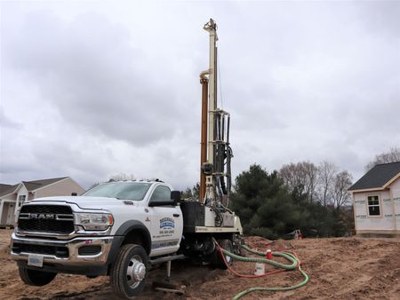 White drilling truck with tall rig on a muddy construction site under cloudy skies