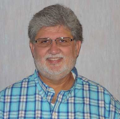 Man with graying hair and beard smiles, wearing glasses and a blue plaid shirt.