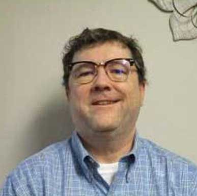 Man wearing glasses, smiling, in blue plaid shirt, indoors.