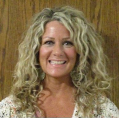 Woman with curly blonde hair smiles at the camera, wearing a white top, against a wood-paneled wall.