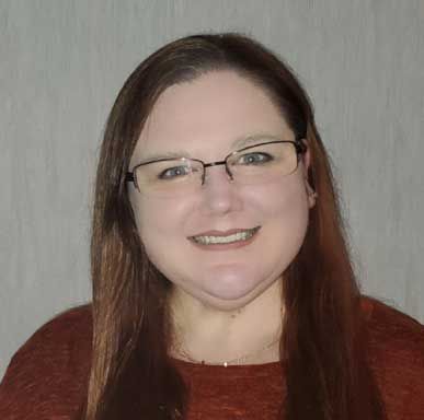 Woman with glasses smiles; long brown hair, wearing a reddish-brown top.