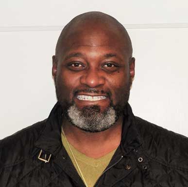 Man with a salt-and-pepper beard smiles, wearing a black jacket and green shirt against a white wall.
