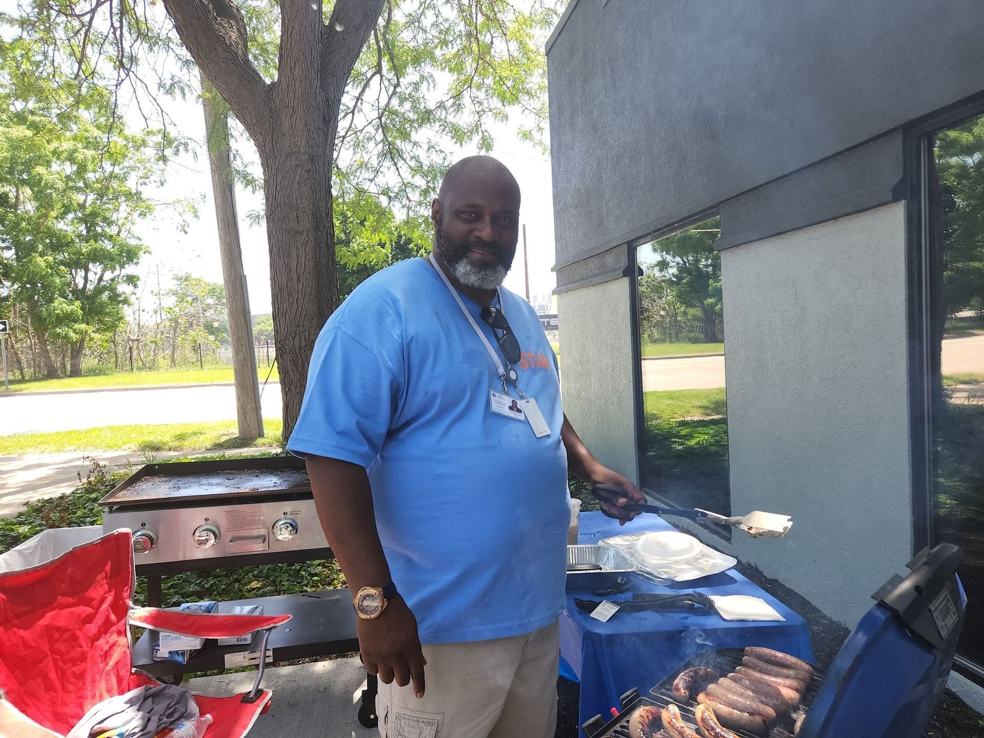 Man grilling food outdoors, smiling. Blue shirt, light pants, next to a grill.