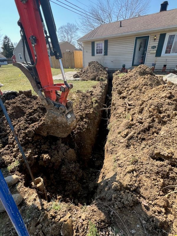 An excavator digging a trench in front of a beige house on a sunny day.