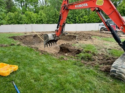 A red Kubota excavator digs a hole in a grassy yard.