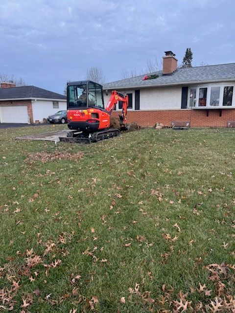 An orange mini excavator on a lawn next to a house, digging. Overcast sky.