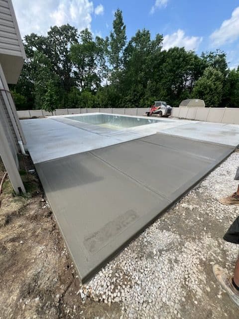Newly poured concrete patio surrounding a pool, construction site. Gray concrete, blue sky, trees in the background.