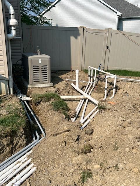 Installation of underground electrical conduit near a home's generator, with white pipes and a brown fence.